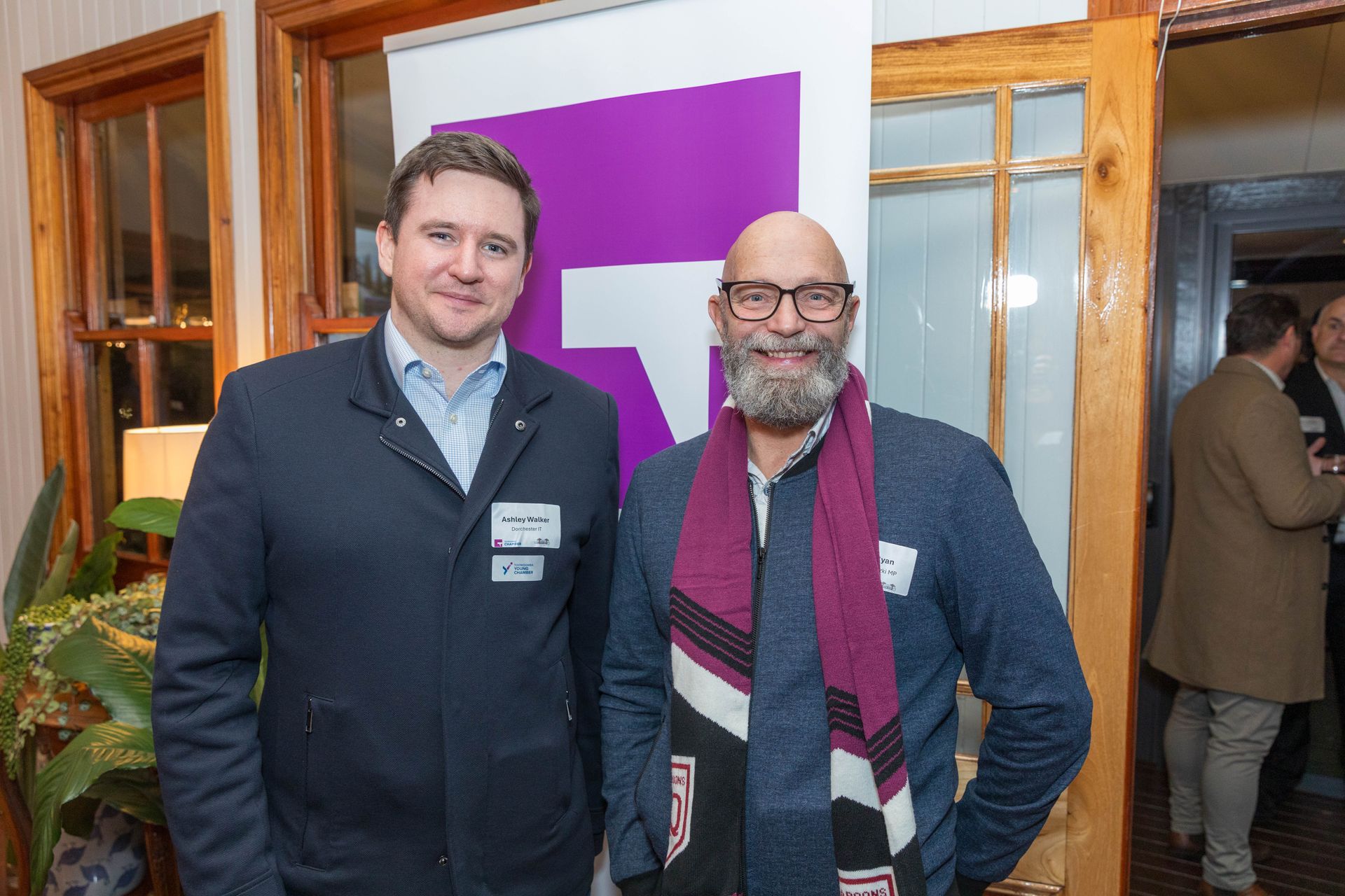 Two men posing together indoors. One wears a dark coat, the other a blue sweater and scarf. A purple banner is behind them.