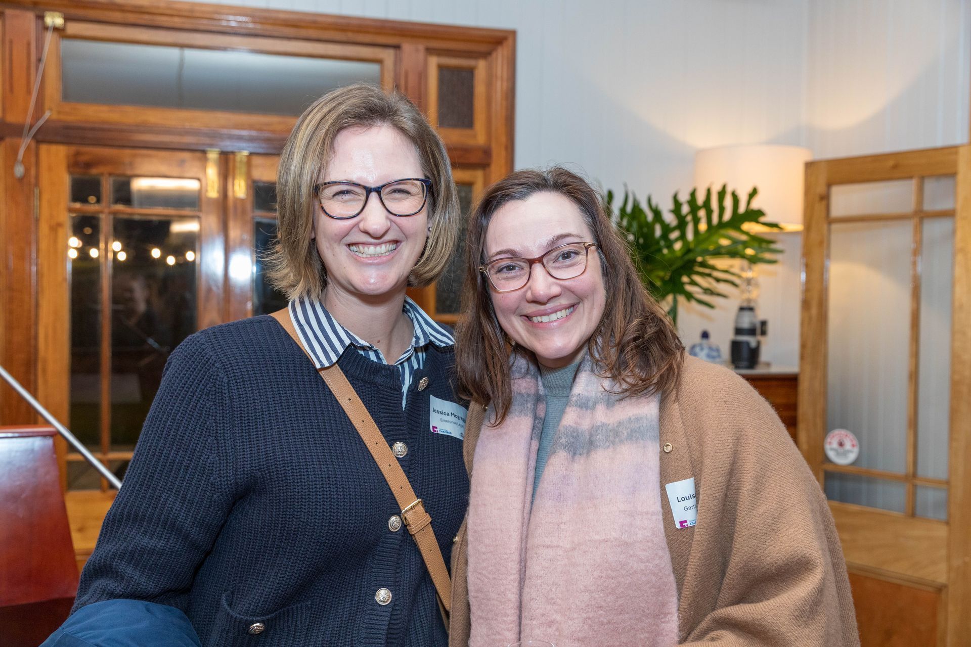 Two women smiling indoors. One wears glasses, a blue sweater over a striped shirt, and a crossbody bag. The other wears glasses and a tan coat with a pink scarf.