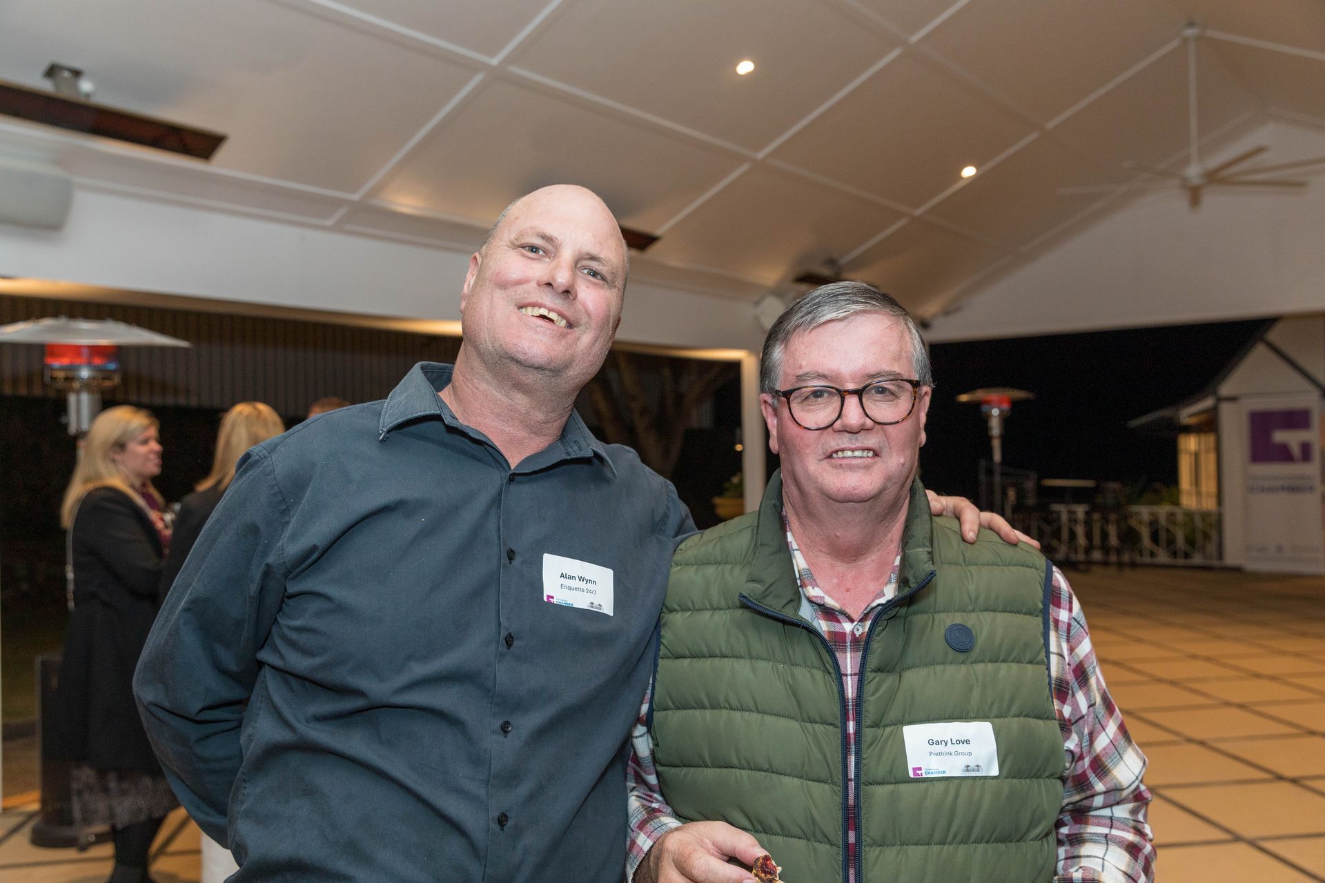 Two men at an outdoor event, smiling and posing for a photo. One wears a green vest, the other a blue shirt.