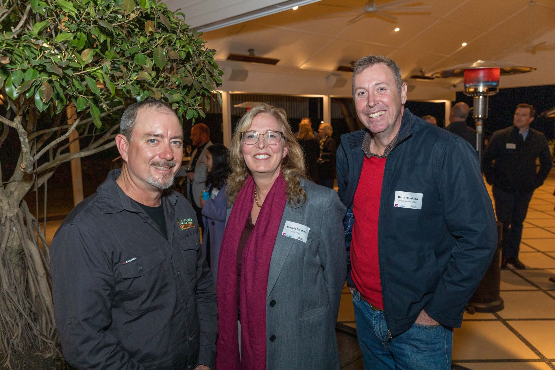 Three people smiling at a gathering. A man in a black shirt stands on the left, a woman in a grey blazer in the middle, and a man in a blue jacket on the right.