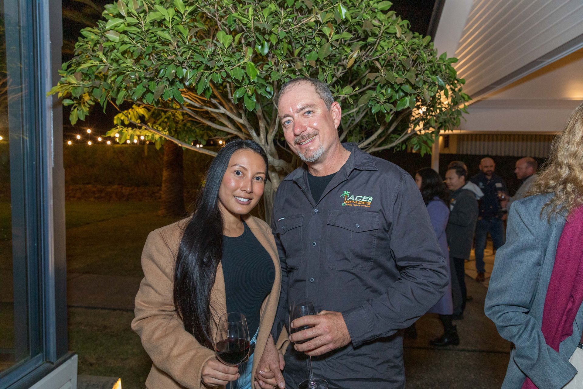 A man and woman smile while holding wine glasses outdoors. They stand near a tree, with a building and other people in the background.