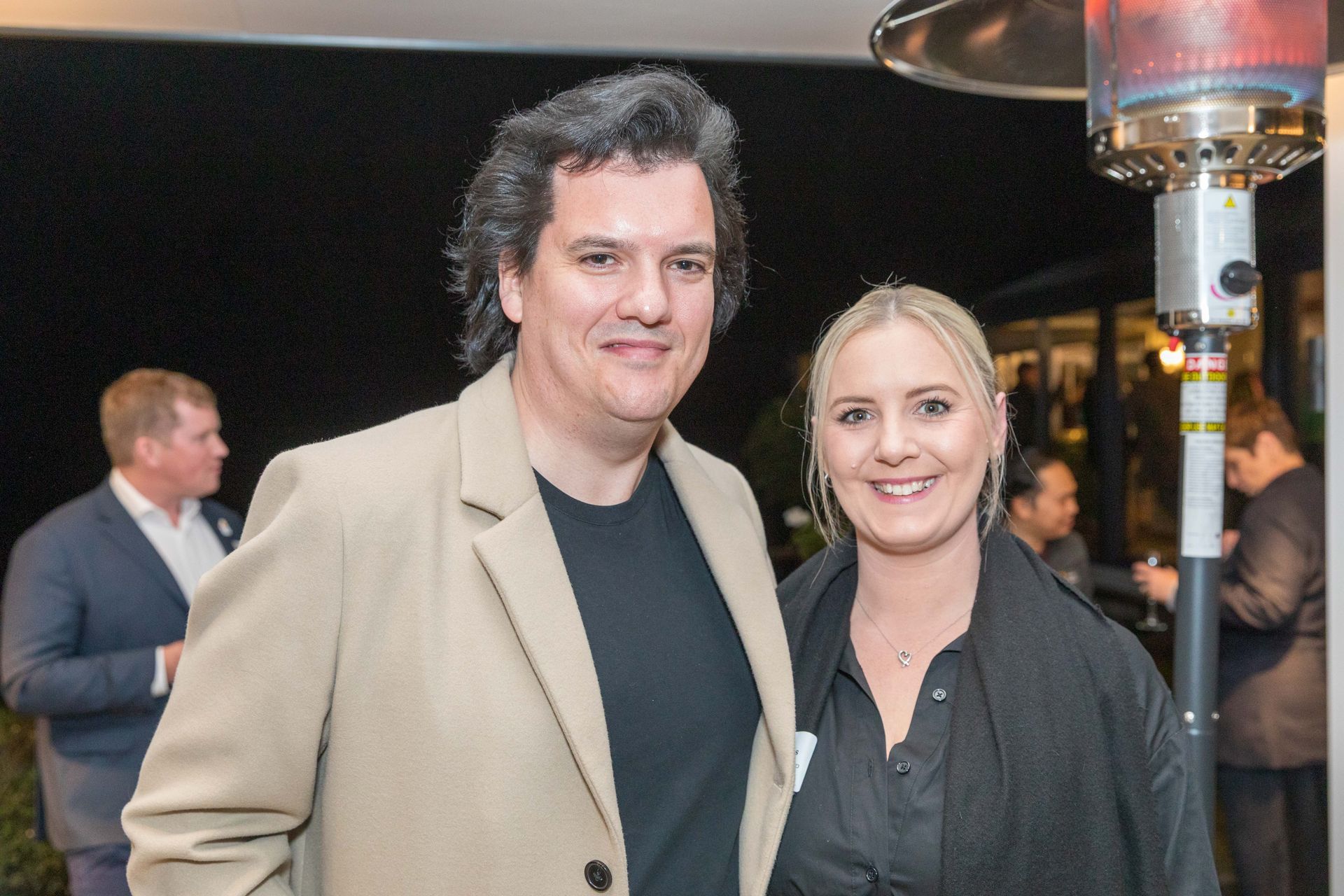 A man and woman smile at the camera, standing outside. The man wears a tan jacket, the woman a black top, at a nighttime event.