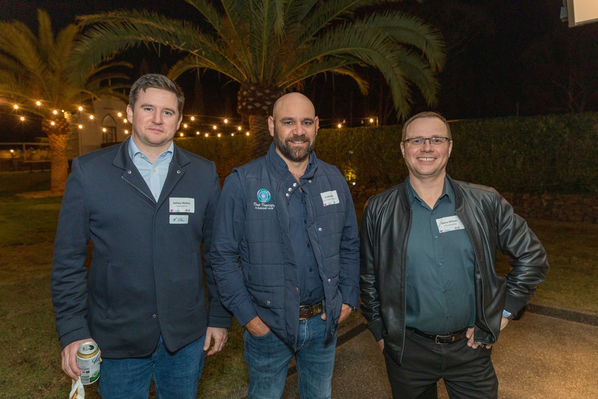 Three men in casual wear posing outdoors at night. They're smiling, in front of palm trees, and string lights.