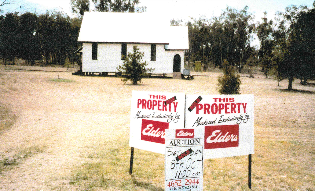 A white church with a sale sign in front, advertised by Elders. The setting is a grassy field with trees.