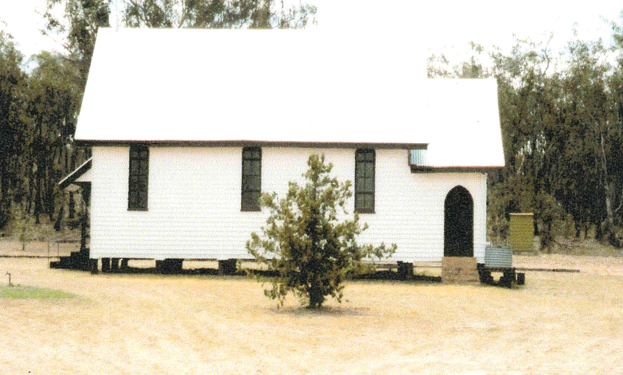 White wooden church with a tree in front, set in a dry, outdoor area.