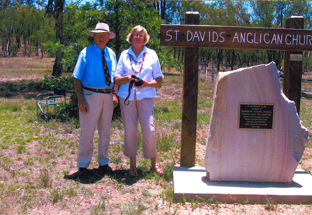 Two people stand by a sign for St. David's Anglican Church. The sign is next to a stone memorial, set in a grassy area.