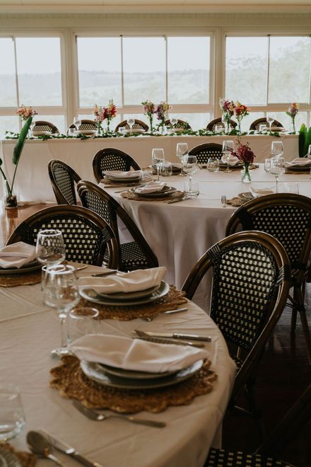 Event hall with rustic decor. Hanging Edison bulbs and floral arrangements over tables set for guests with white chairs.