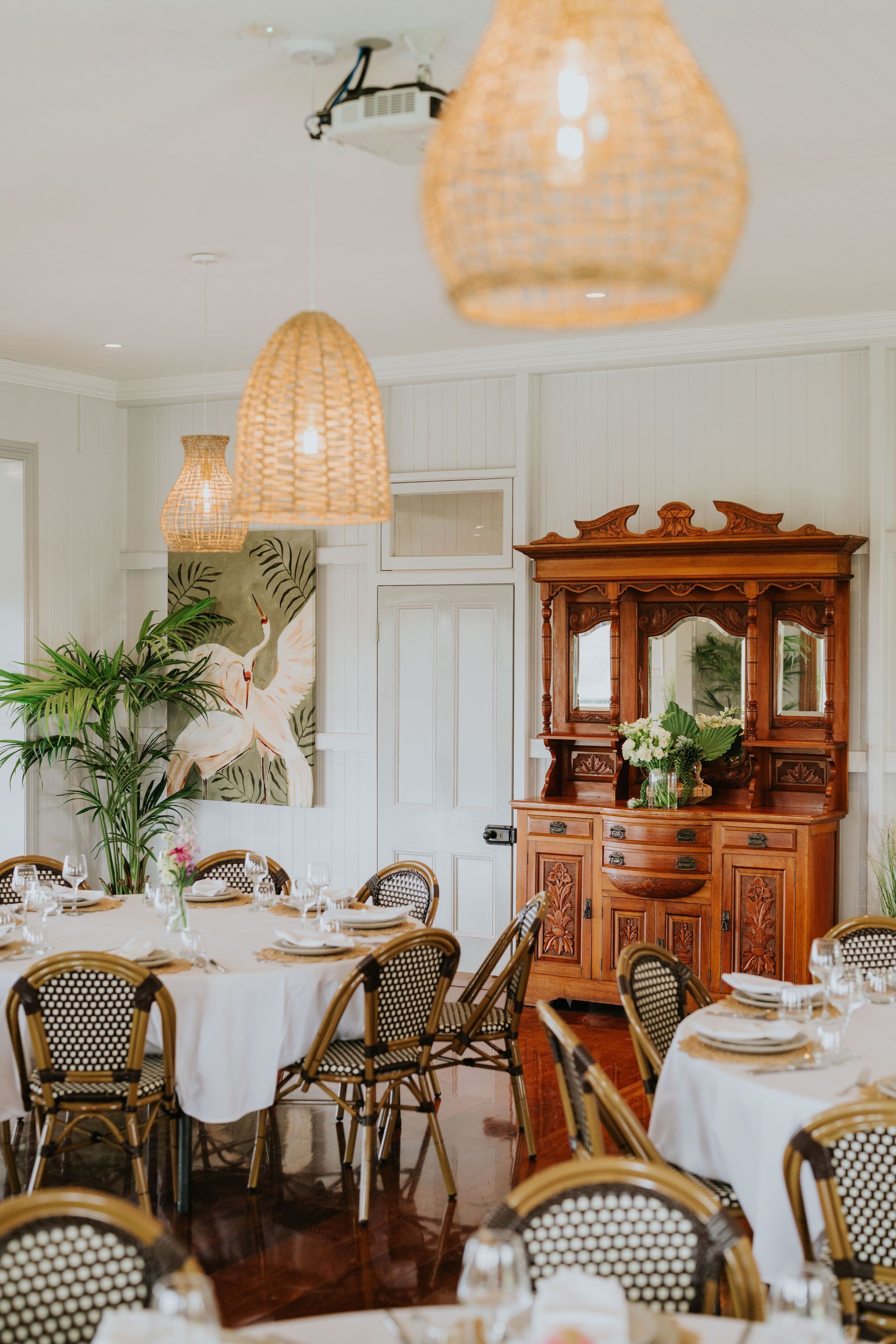 Event hall with rustic decor. Hanging Edison bulbs and floral arrangements over tables set for guests with white chairs.