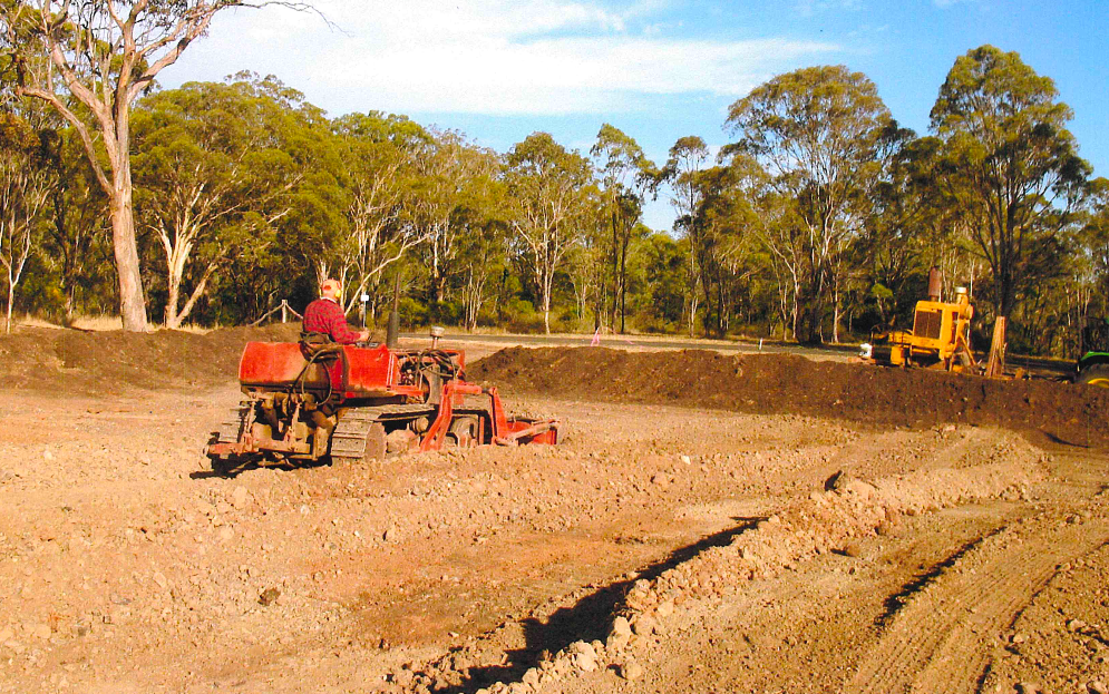 A person operates a small tractor tilling soil in an open, brown field, with trees and a yellow bulldozer in the background.