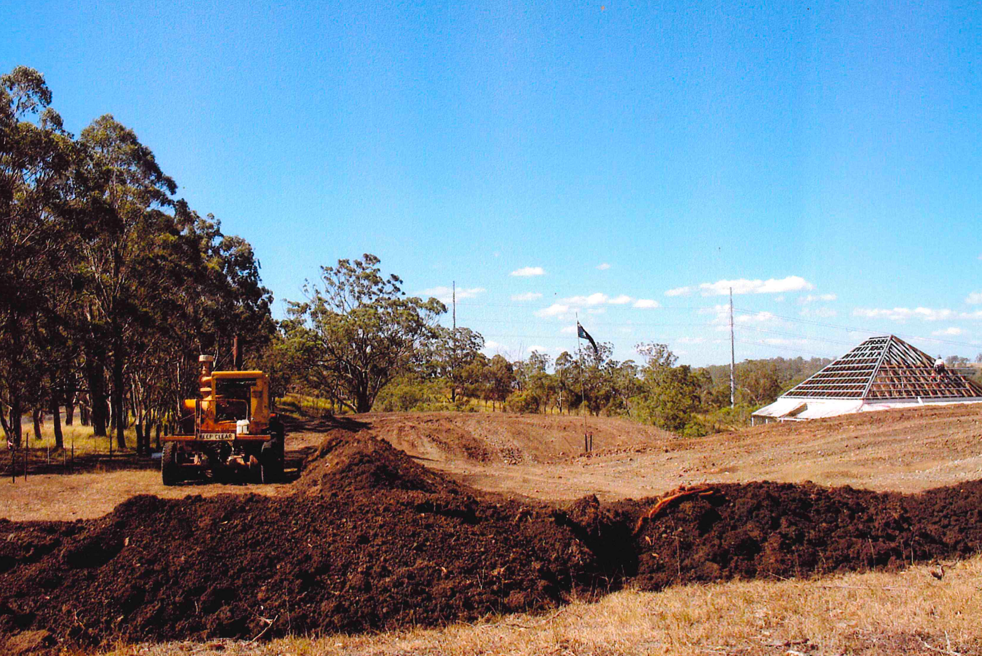 A yellow bulldozer piles dark soil in a field near trees and a building under construction, under a blue sky.