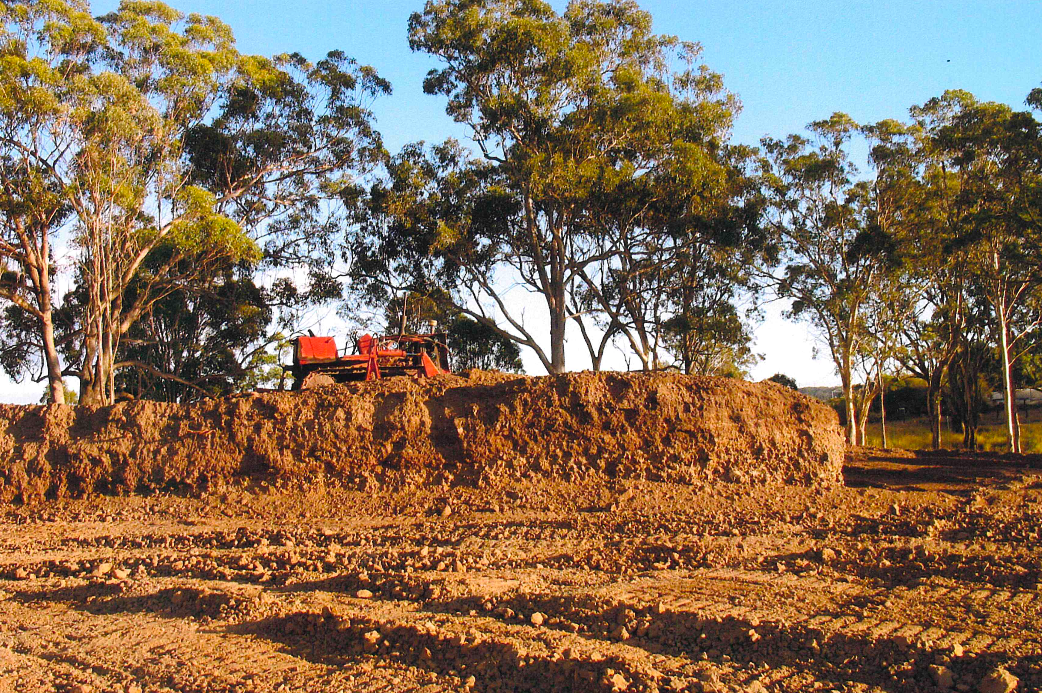 A tractor works on a large pile of brown material in a field, trees in the background under a blue sky.