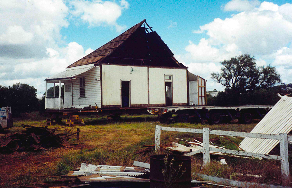 Old, white house with a damaged roof and missing siding, sitting on beams in a field. Debris and a white fence are in the foreground.