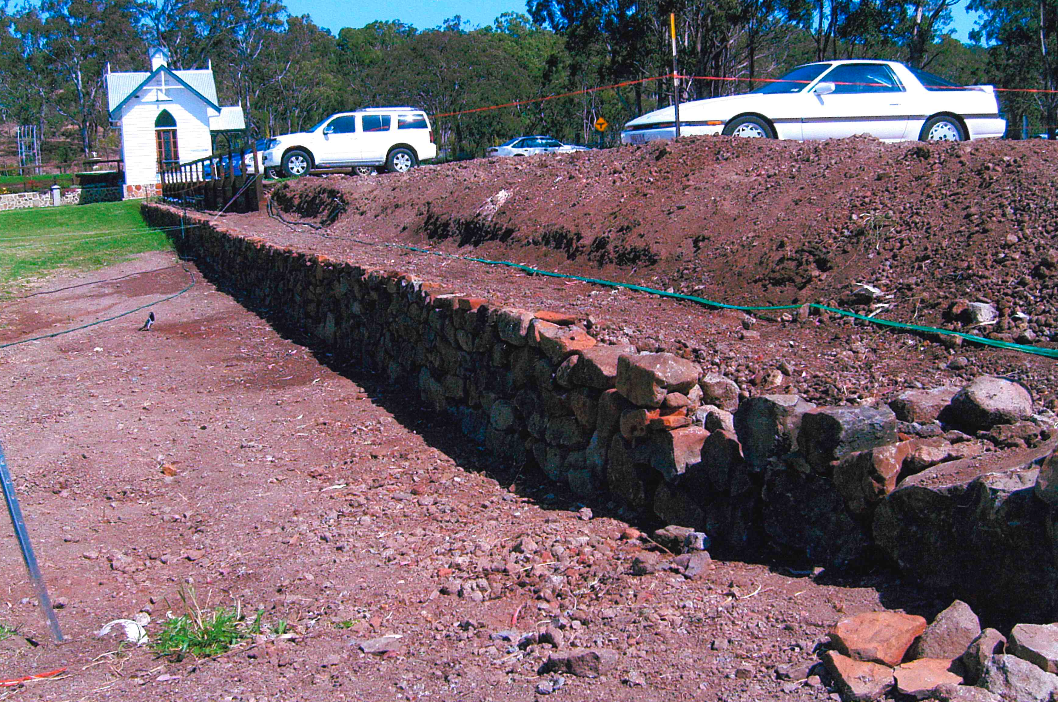 A long, low brick wall under construction, next to a dirt path and a small white chapel. Cars are parked on a slight rise in the background.