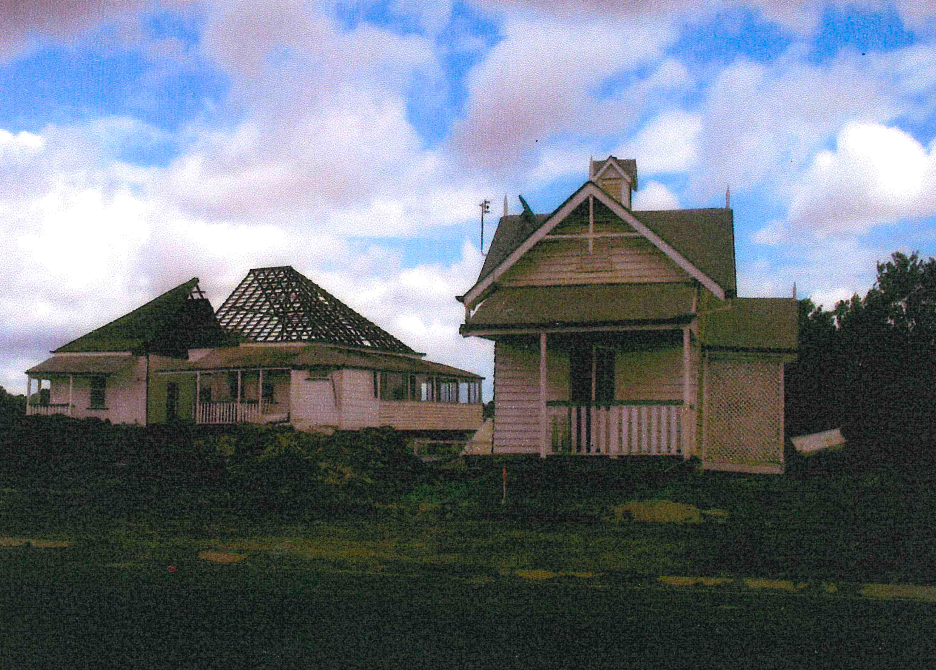 Two white houses with angled roofs sit on a grassy area under a cloudy sky. One house is missing a roof.