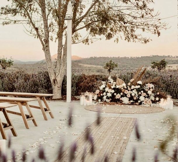 Wedding ceremony setup in a lavender field with a flower-covered altar and wooden benches facing a scenic valley at sunset.