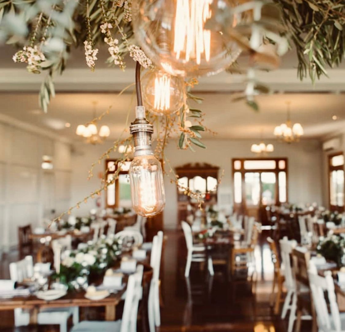 Event hall with rustic decor. Hanging Edison bulbs and floral arrangements over tables set for guests with white chairs.