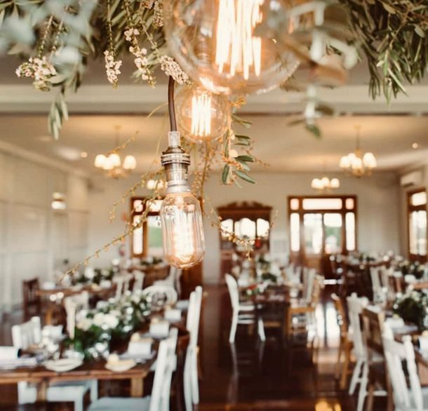 Event hall with rustic decor. Hanging Edison bulbs and floral arrangements over tables set for guests with white chairs.