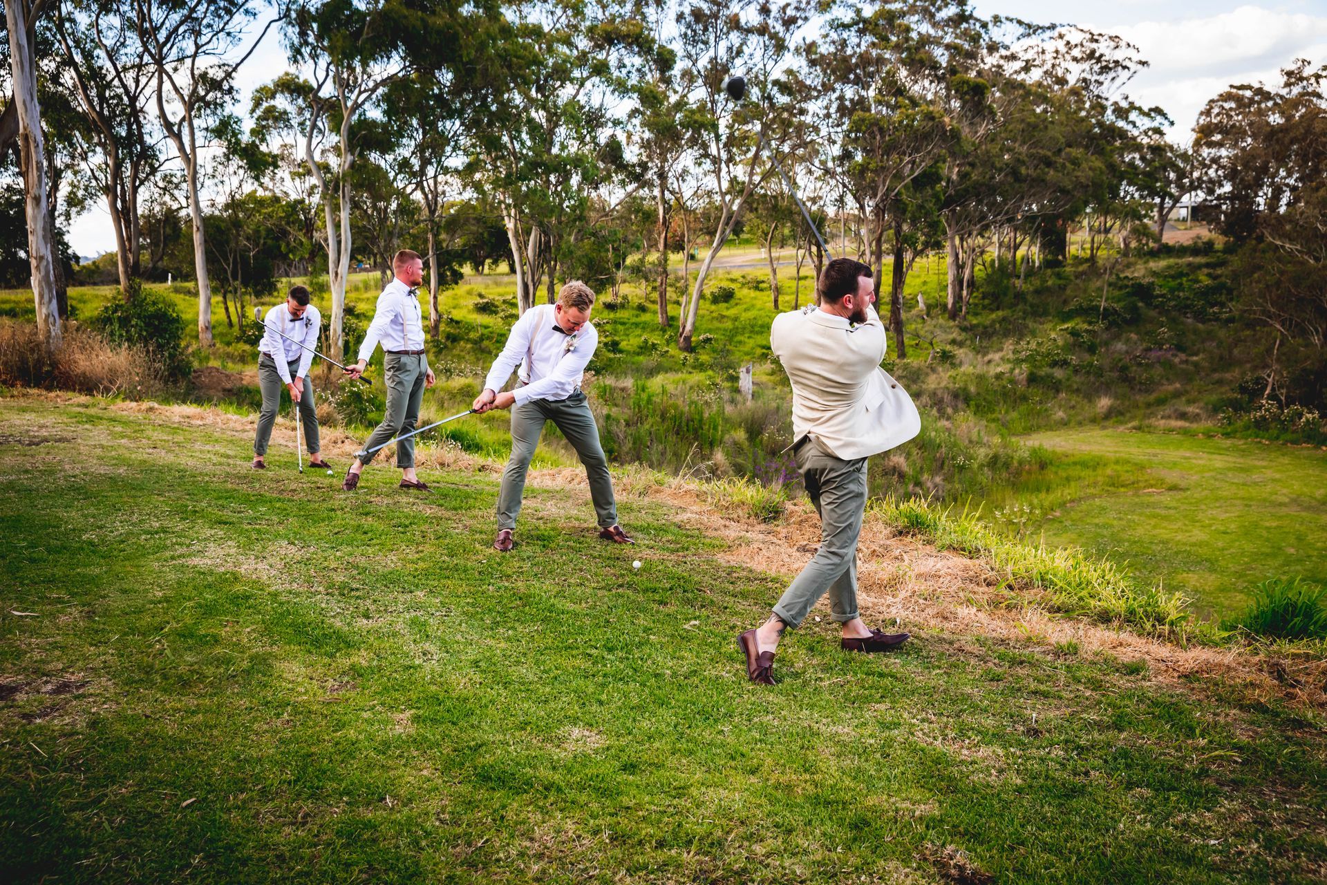Men, in matching outfits, swinging imaginary golf clubs on a grassy hill. Sunlight, trees, and a natural setting.