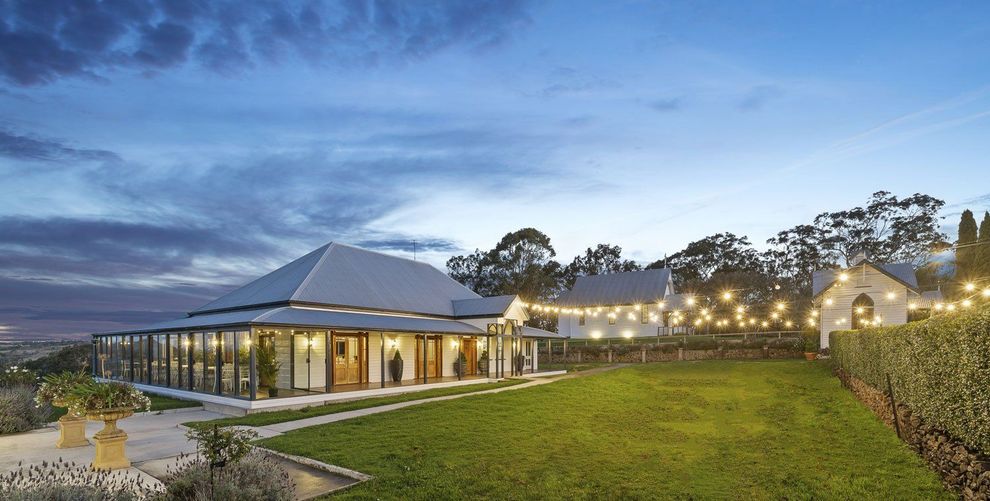 A dusk view of a large event venue with a long veranda, string lights, and a manicured lawn under a cloudy sky.