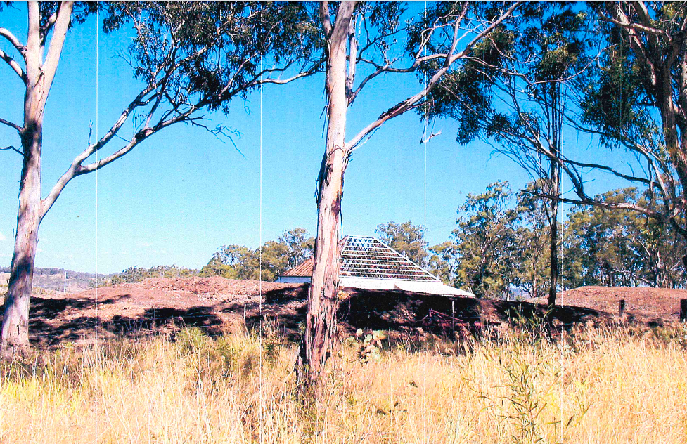 A building with a tall antenna is visible amidst trees and dry grass, under a clear blue sky.