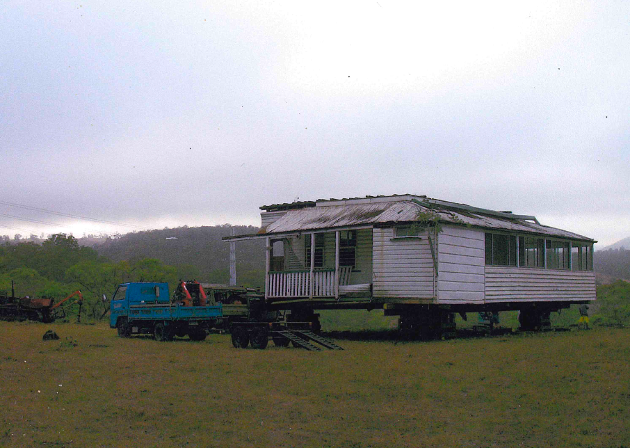 An old, white, weathered house on wheels is being towed by a blue truck in a grassy field. The sky is overcast.