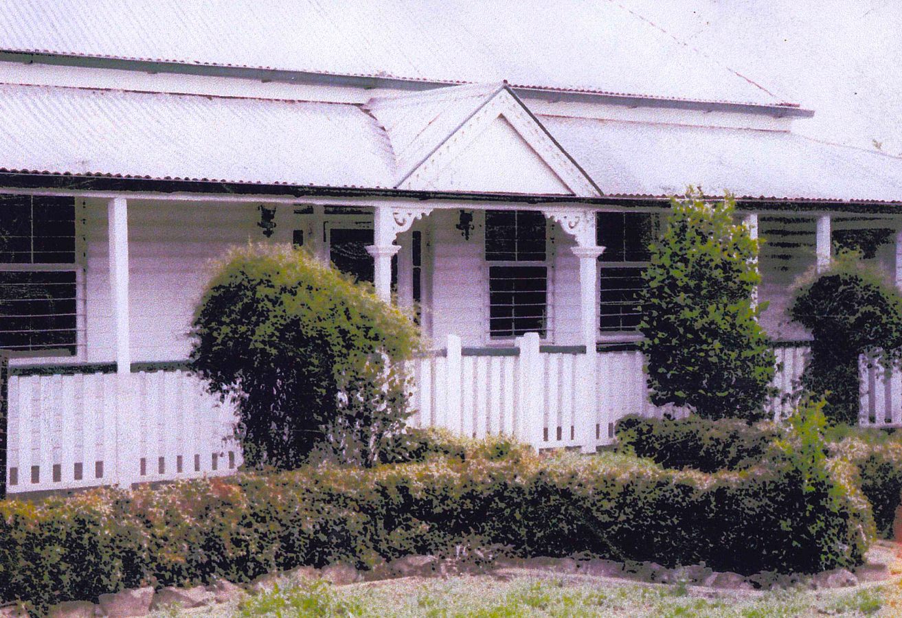 White house with a porch and corrugated iron roof. Bushes and shrubs are in front of the house.
