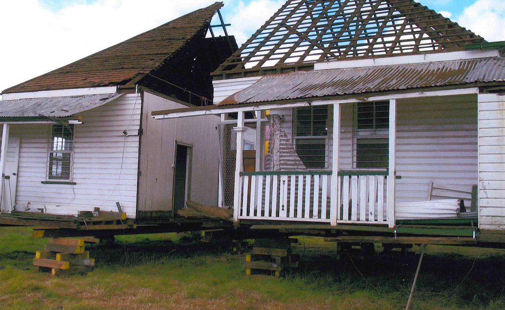 Dilapidated white house with a damaged roof, sitting on wooden supports in a grassy yard. The roof is partially missing, exposing the frame.