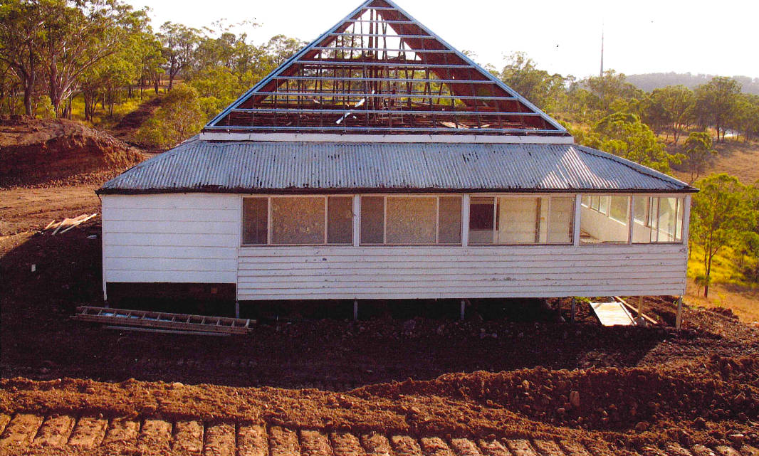 White house with a partially collapsed pyramid-shaped roof, on a dirt mound. The house has windows and sits in a rural setting.