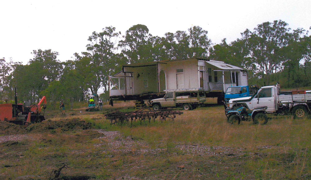 A house being transported on trucks in a grassy field, with construction equipment and vehicles nearby. Overcast sky and trees in the background.