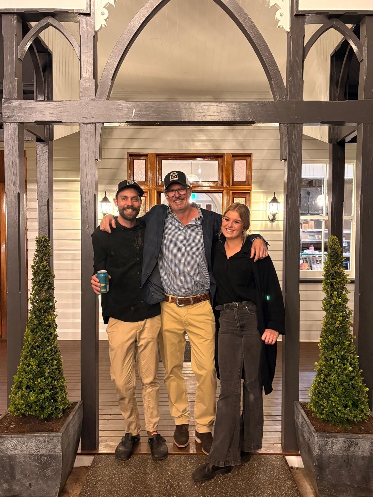 Three people pose under a wooden archway; all smiling. 