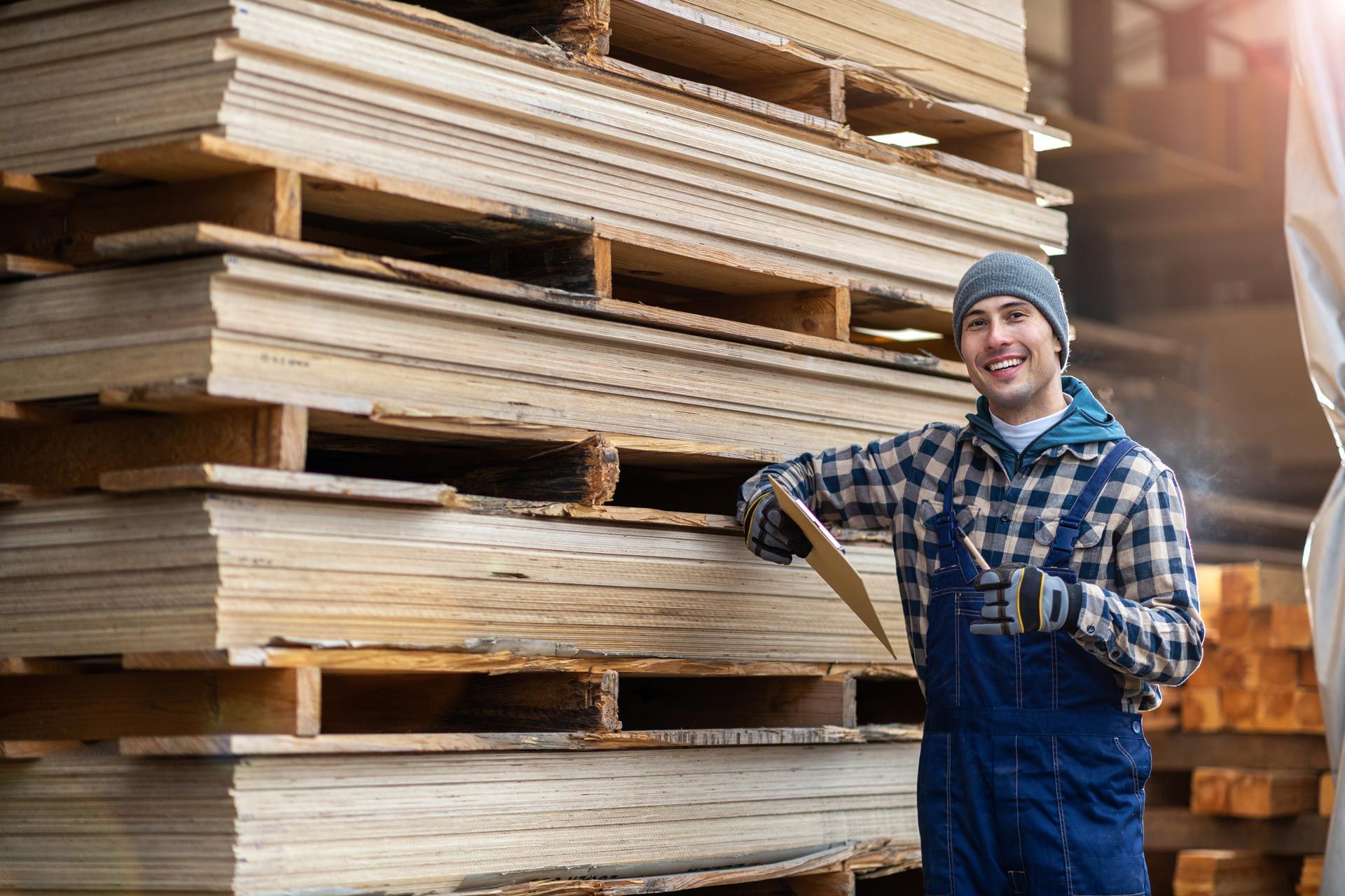 Worker in overalls holding clipboard beside stacked lumber in a warehouse.