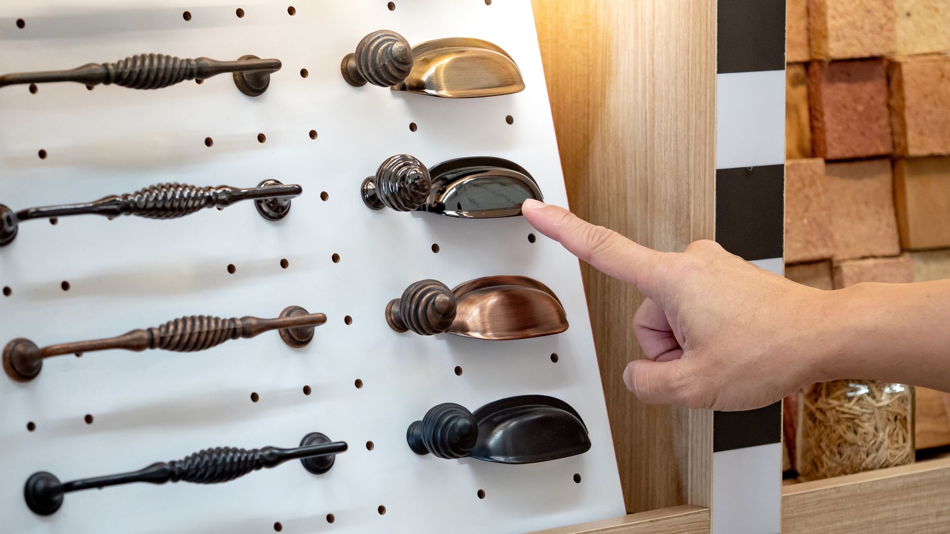 A hand pointing at a cabinet handle on a display board with decorative handles arranged in rows. A hand pointing at a cabinet handle on a display board with decorative handles arranged in rows.