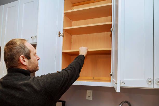 Man inspecting empty white kitchen cabinets with wood shelves during home renovation or remodel.