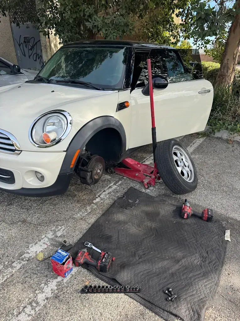 White Mini Cooper with a flat tire, jacked up; tools and spare tire nearby.