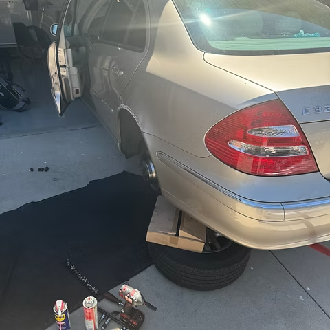 A gold Mercedes-Benz, rear wheel removed, supported by a tire and wooden block, parked in a driveway.