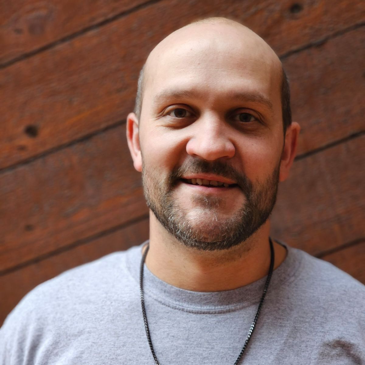 Man with a bald head and short beard smiling at the camera, wearing a gray shirt, in front of wood paneling.