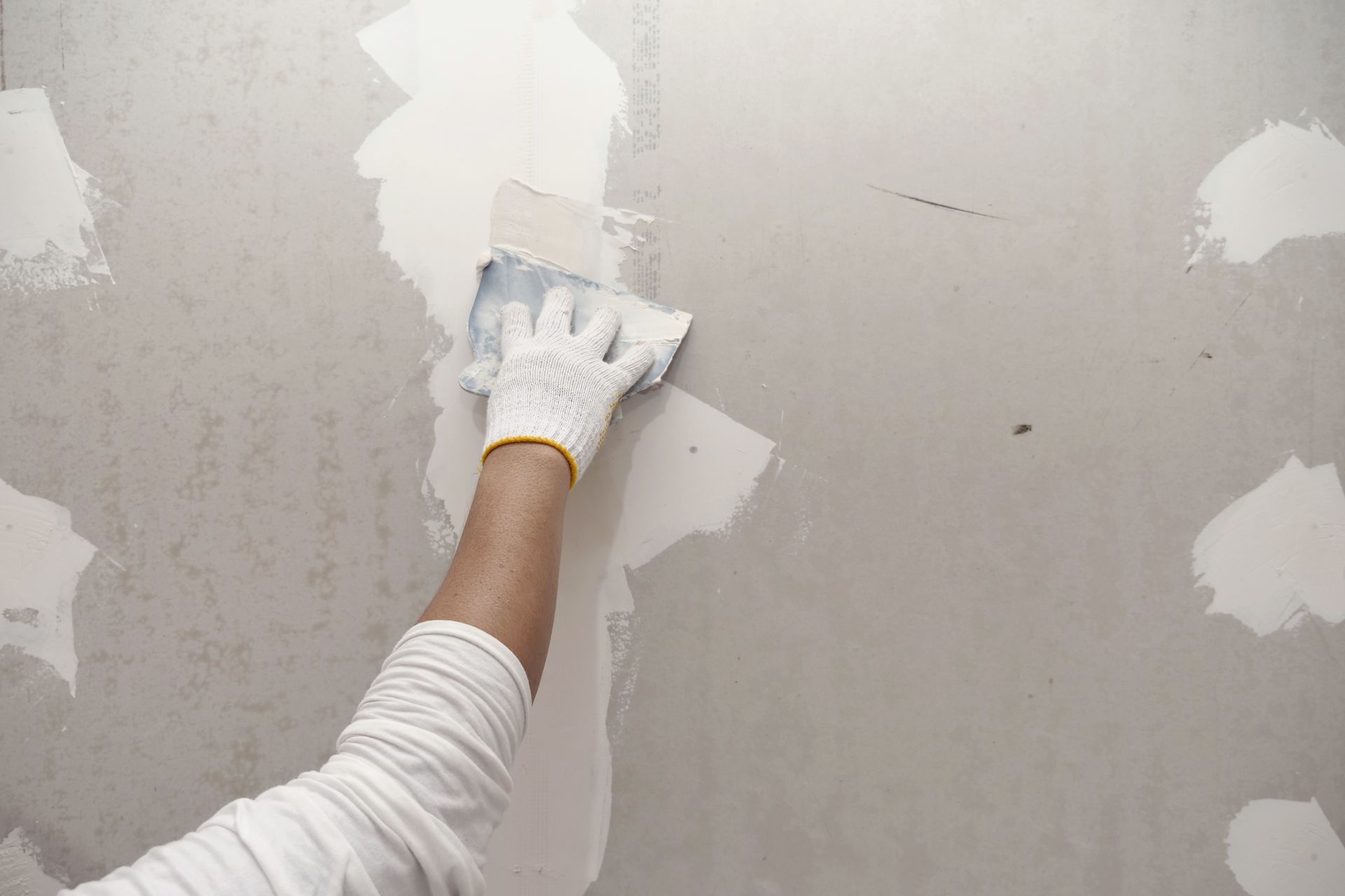 A person wearing a white glove uses a putty knife to apply joint compound to a gray drywall surface.