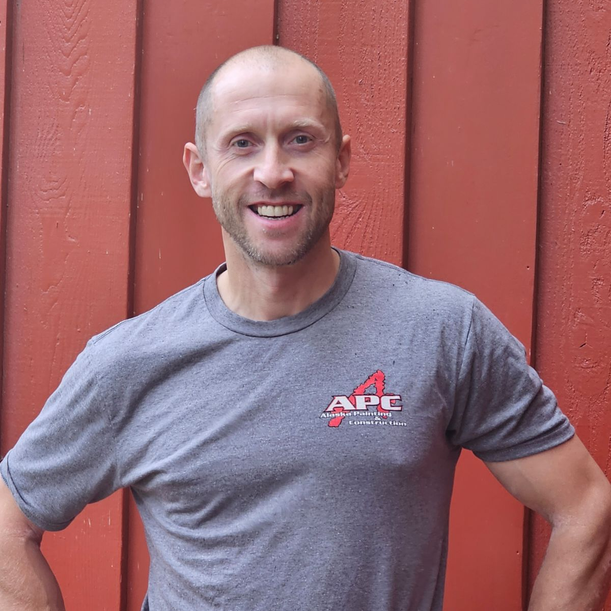 Man with shaved head wearing a gray shirt, smiling in front of a red wooden wall.