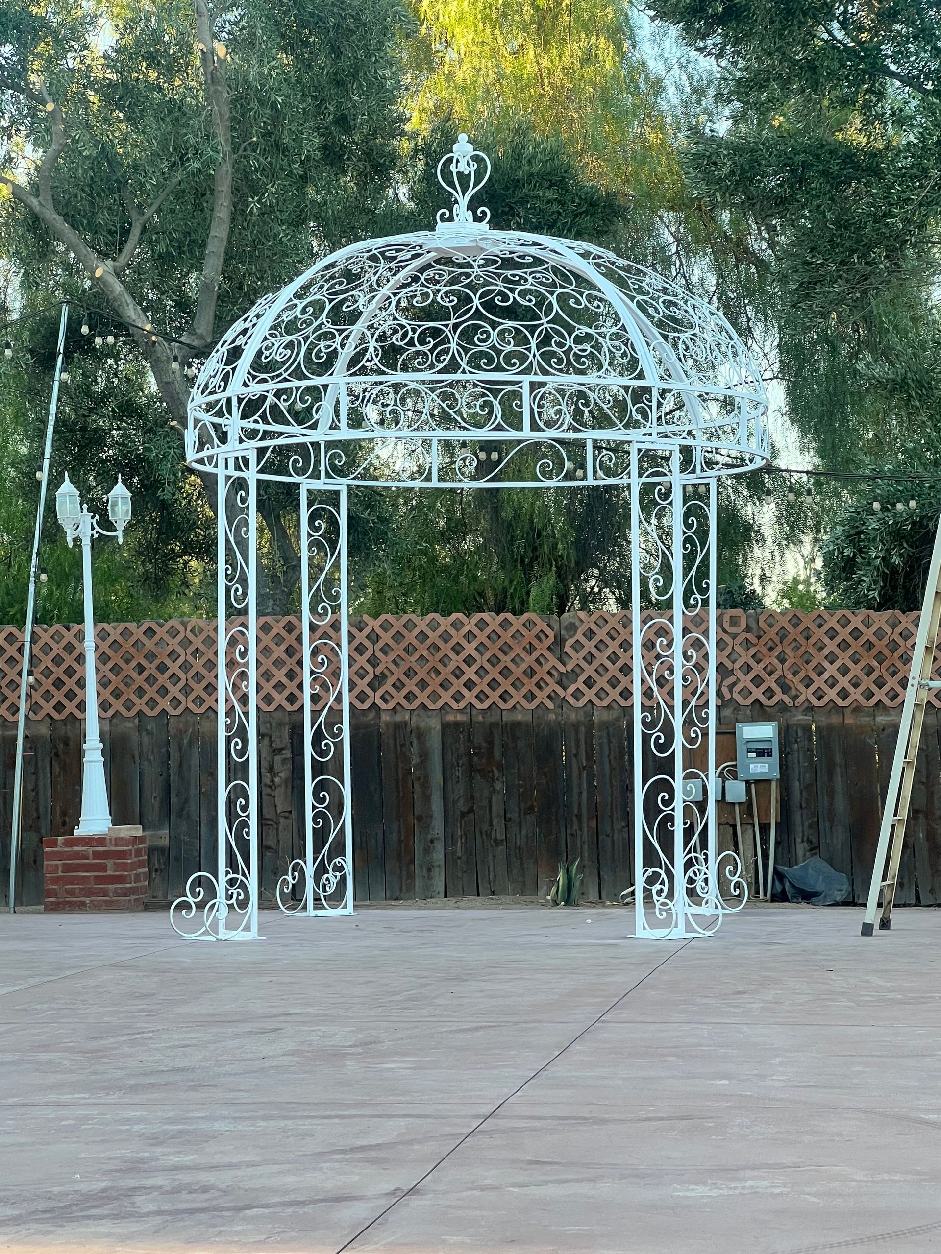 A white gazebo is sitting on a brick pavement in front of a wooden fence.