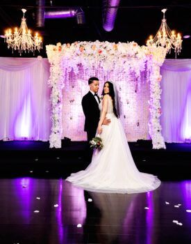 A bride and groom are standing on a stage in front of a floral arch.