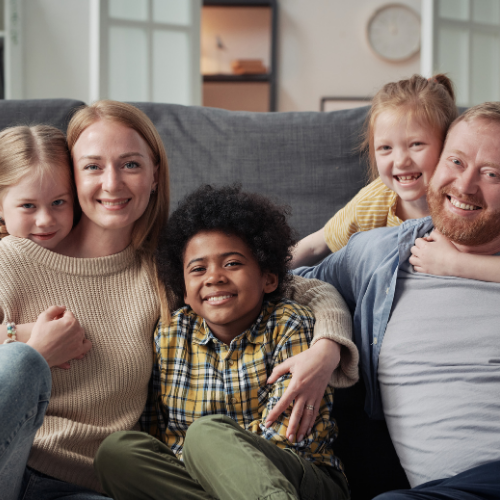 Family of five smiles together on a couch, indoors.