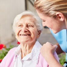 Smiling elderly woman, in pink, with a caregiver in blue, outside with flowers.
