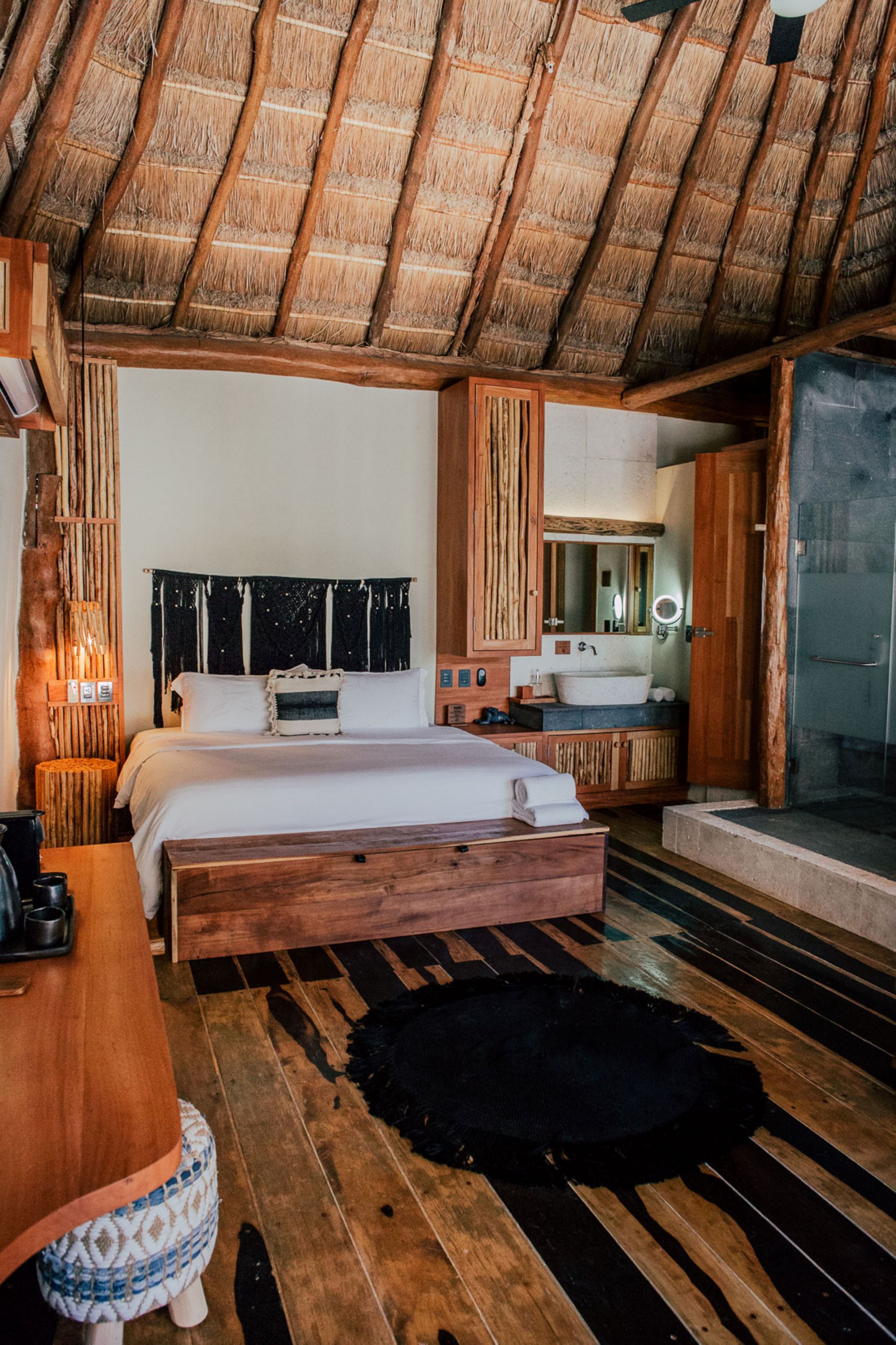 Bedroom with wooden furniture and thatched roof. White bedding, black rug, and bathroom with glass door.