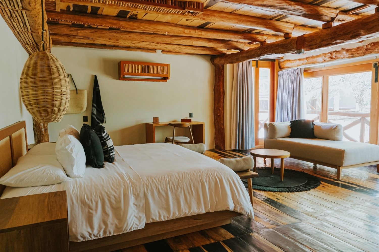 Bedroom with wooden beams, a bed, a couch, and a woven hanging basket, in a rustic setting.