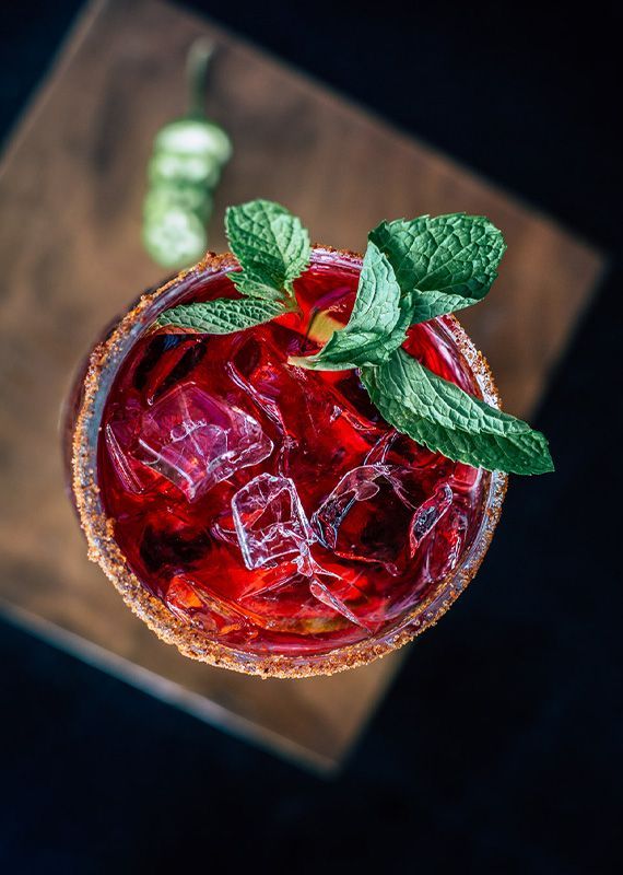 Red cocktail with ice, mint, and a chili pepper on a wooden board.