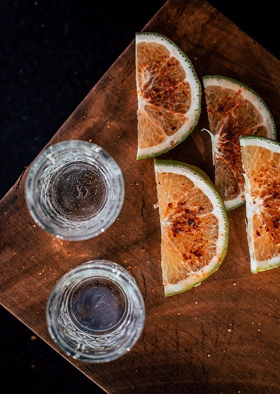 Two clear shot glasses and orange slices on a wooden cutting board, with a dark background.
