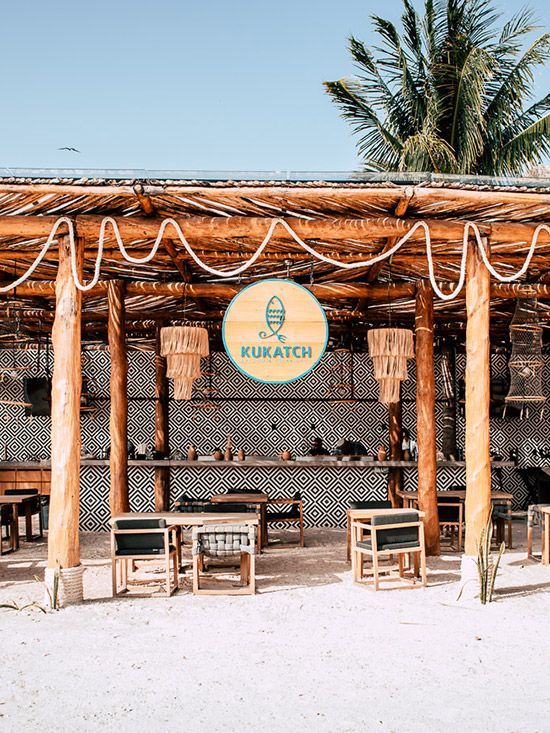 Beachside restaurant Kukatch with a thatched roof, wooden furniture, and circular sign.