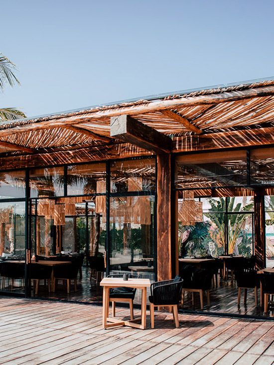 Restaurant with wooden beams and thatch roof. Tables and chairs visible inside. Blue sky.