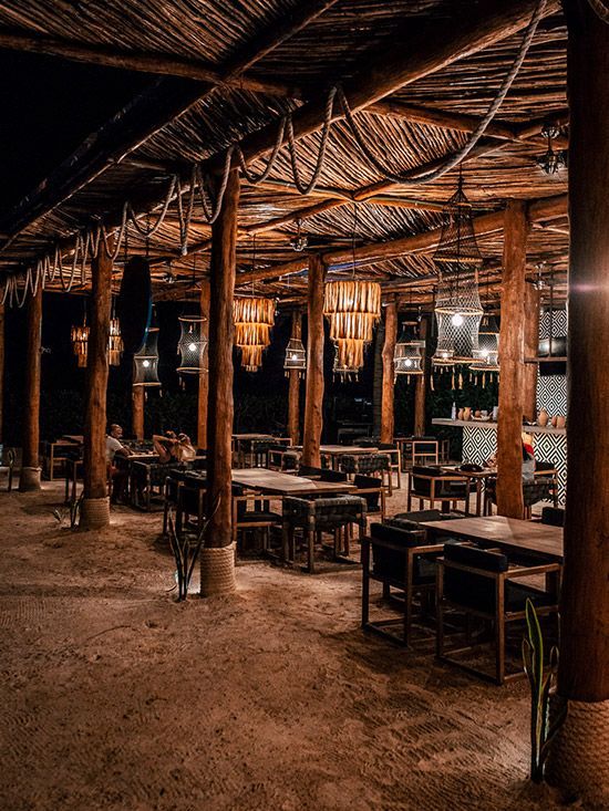 Restaurant with wooden posts, thatched roof, tables, chairs, and hanging lights at night.