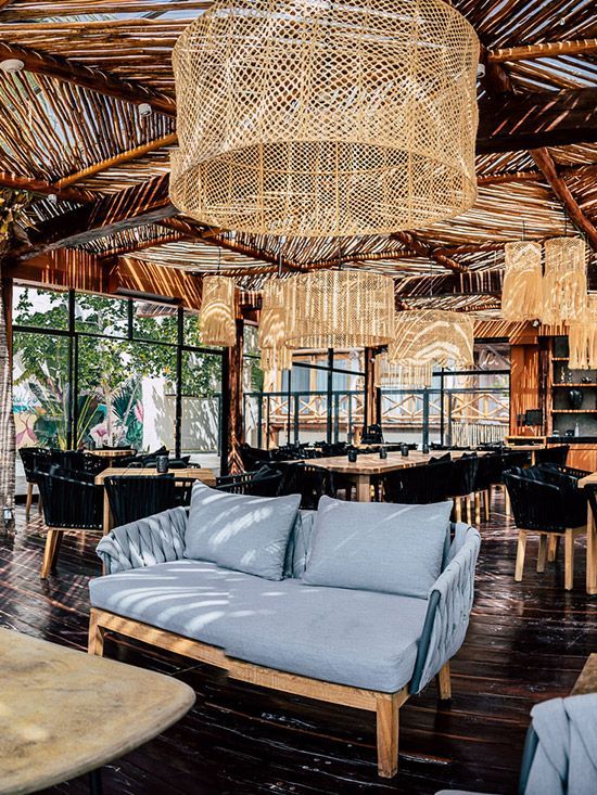 Restaurant interior: woven lamps, wooden beams, gray sofa, tables, chairs, and dark floor.